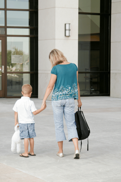 Mother dropping young boy off at preschool. She is holding the boy's hand and carrying a black backpack in the other hand. Both of their backs are to the camera.