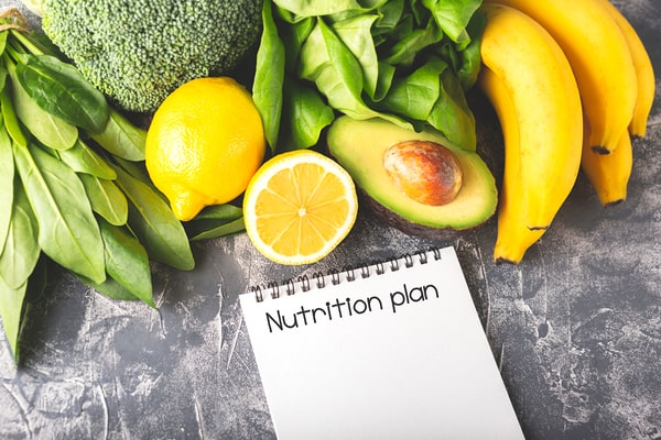 "Nutrition plan" written on a notebook, alongside a slice of avocado, banana, lemon, and vegetables