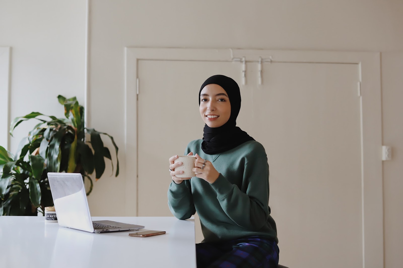 A young woman wearing a hijab drinking coffee in her office and smiling