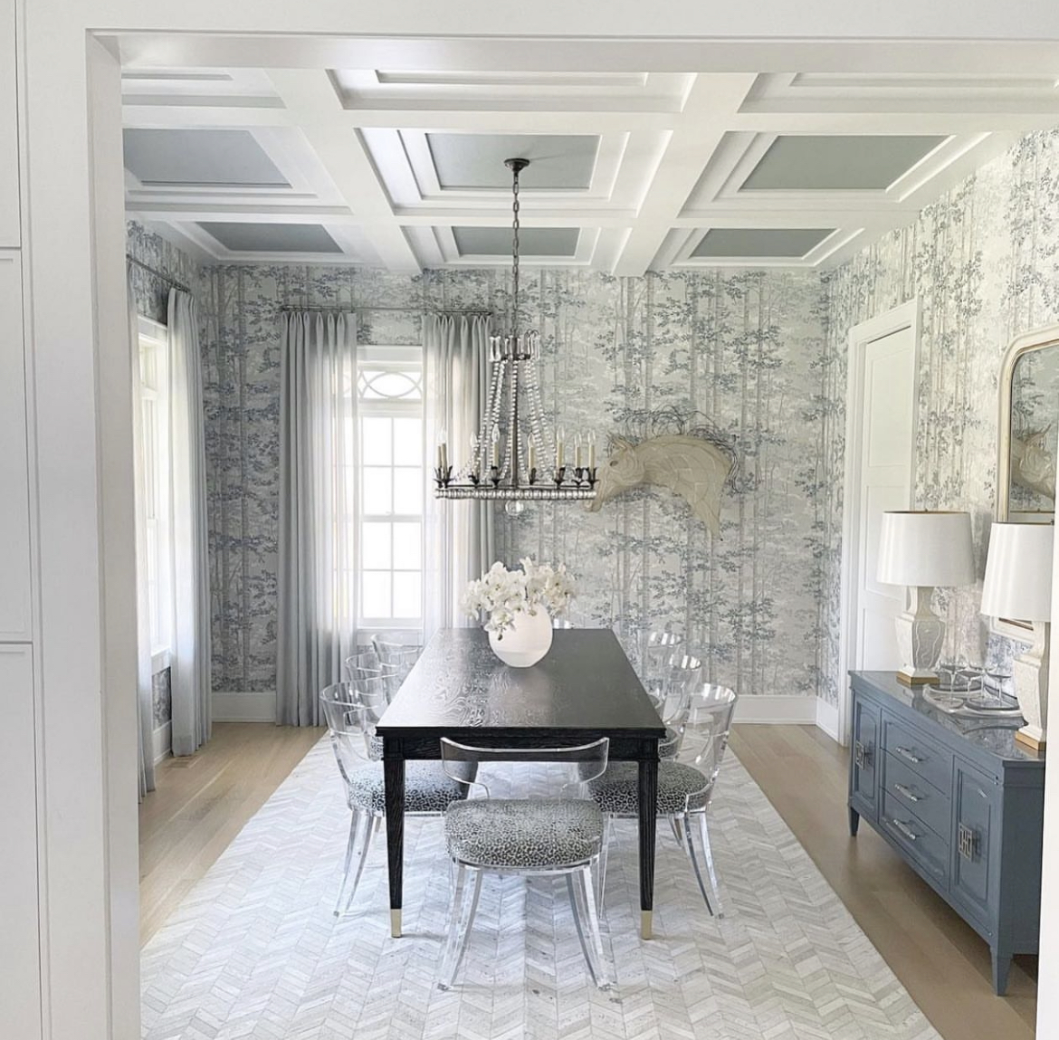 Blue and white tones dominate the color scheme in this dining room, illuminated by a stunning chandelier that hangs above a wooden table that is flanked with modern clear, acrylic chairs.A set of sleek acrylic chairs with comfy cushions surround the table, offering both comfort and modern flair. The chairs provide a striking contrast against the wooden table, creating a visually appealing contrast.A beautiful blue cabinet adds a pop of color to the room while also offering additional storage for dinnerware and other dining essentials. The cabinet complements the blue hues found throughout the dining room, creating a cohesive and harmonious look.The walls are covered in a stylish wallpaper that features intricate patterns and shades of blue and white. The wallpaper adds texture and depth to the room, creating an immersive atmosphere that is both elegant and sophisticated.This dining room is perfect for entertaining guests, with a combination of modern and classic elements that create a welcoming and visually pleasing space. The overall effect is a space that is both functional and beautiful, allowing you to enjoy meals with family and friends in style.