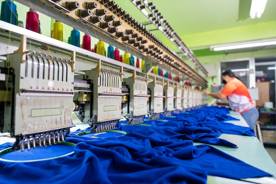 A worker operating a sewing machine in the embroidery area of a textile factory with modern machinery