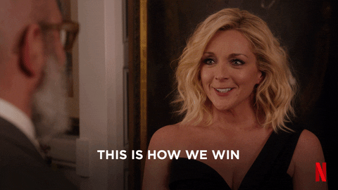 This gif is a scene from Unbreakable Kimmy Schmidt. In this scene, a blonde woman and bald, bearded man wiggle there fingers together in a handshake manner. In front of them is the phrase "THIS IS HOW WE WIN".