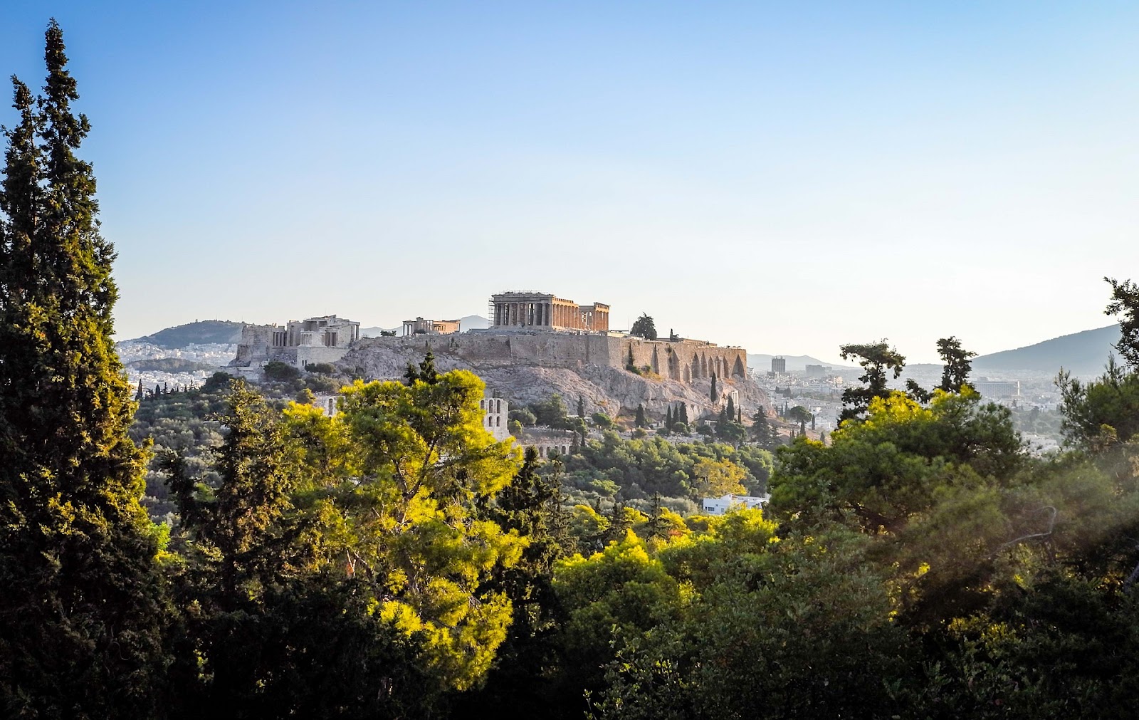 acropolis athens monumental ancient parthenon, seen behind green trees and athens skyline on a clear day.