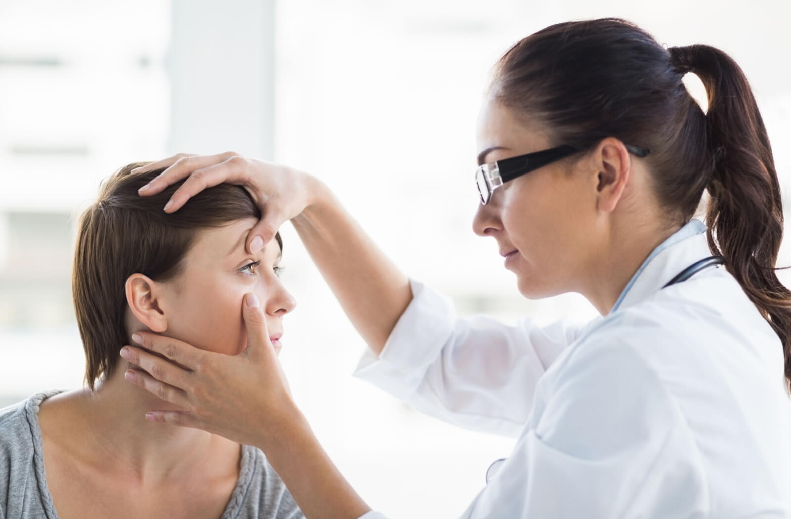 A female doctor is closely examining the patient's eye. The doctor is touching the female patient's right upper eyelid with her right hand and her left hand touching her cheek.