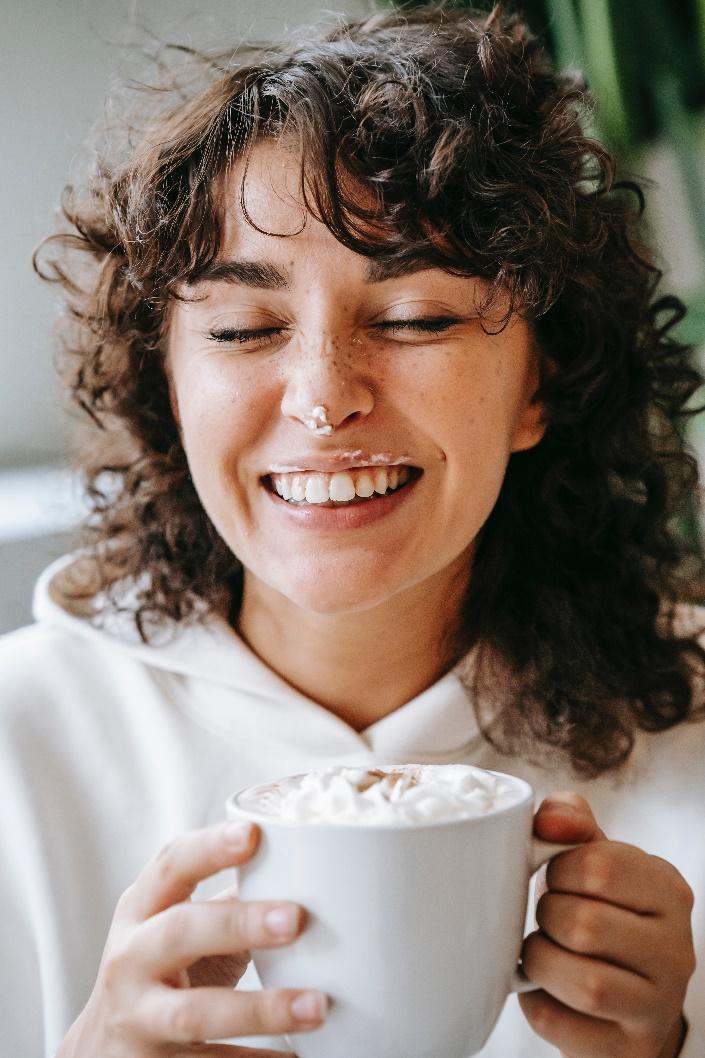 a woman smiling with some whipped cream on her nose while she holds a white mug of a warm drink topped with whipped cream
