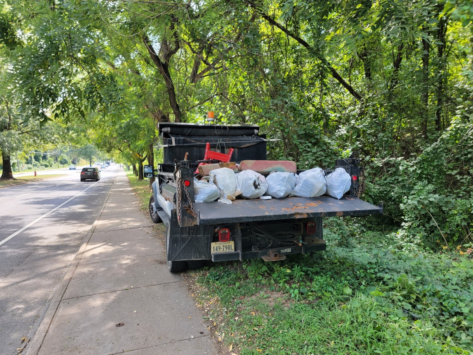 A large city-owned pickup truck is in the center of the photo, with a street and sidewalk to the left and trees, shrubs, and other vegetation to the right. The truck bed is down, and it’s full of half a dozen large plastic trash bags, pieces of a moldy couch, a wooden chair, and whatever is piled beneath them.
