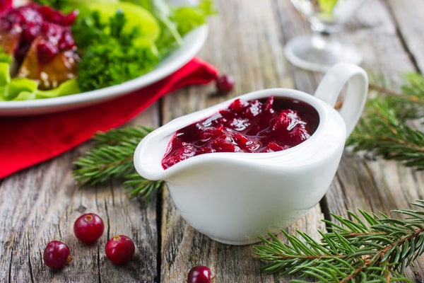 A teacup of cranberry sauce with herbs and a plate of vegetables on the side, placed on a wooden table