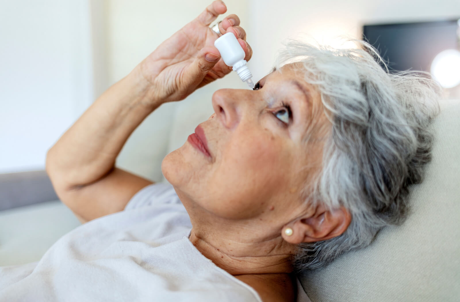 A senior woman leaning back to apply eye drops on her right eye.