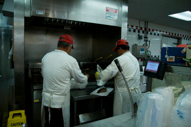 Two chefs in white aprons and red caps stand in front of a stove. They appear to be in a diner. 