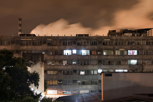A imagem mostra um complexo residencial cinza escuro. Ao fundo, é possível ver o céu alaranjado com uma grande nuvem de fumaça proveniente da usina da cidade.