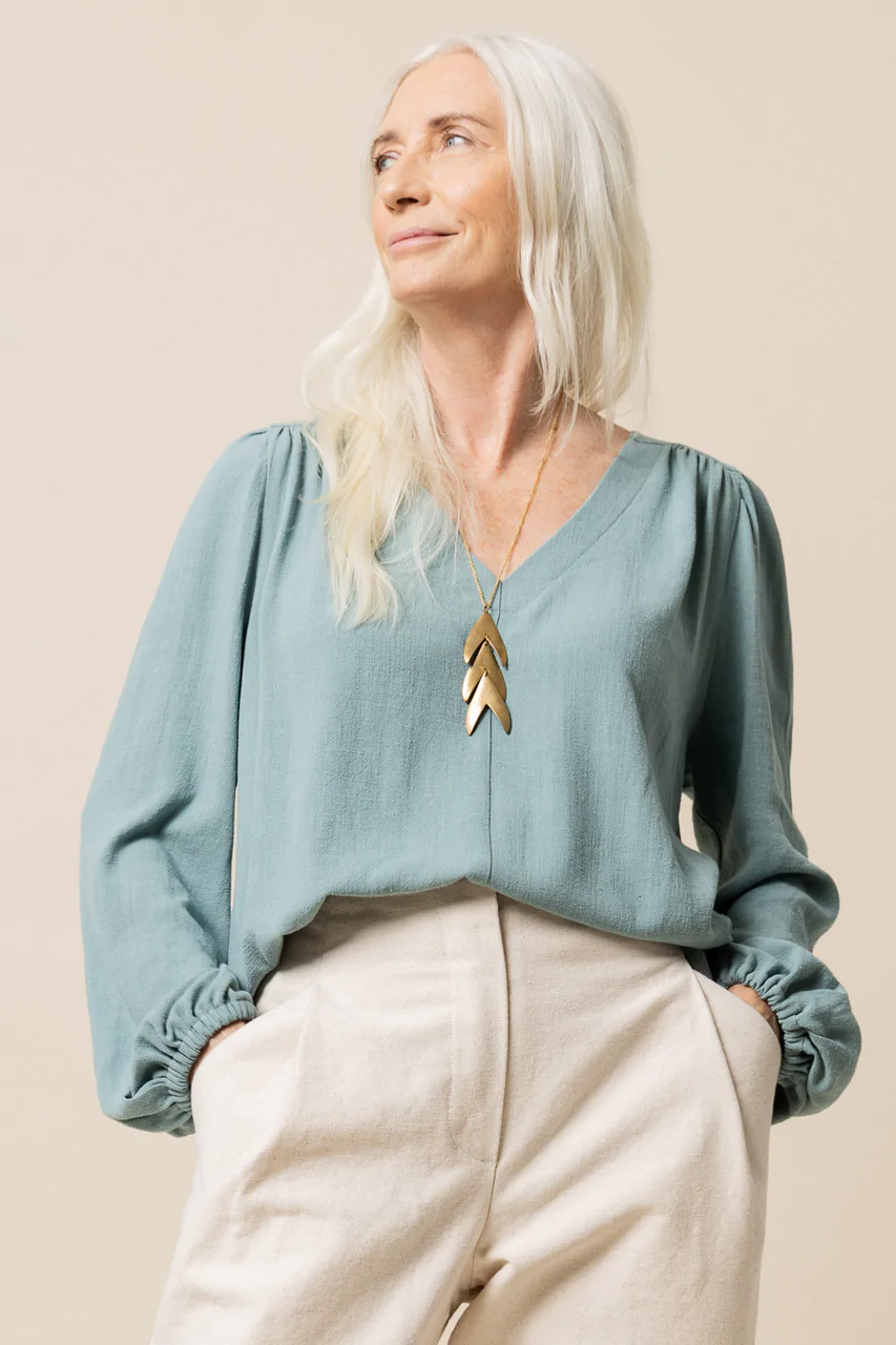 A person with long grey hair is wearing a v-neck top with long full sleeves that are gathered at the wrist with elastic.  The shirt is work with cream pants & a bronze necklace.
