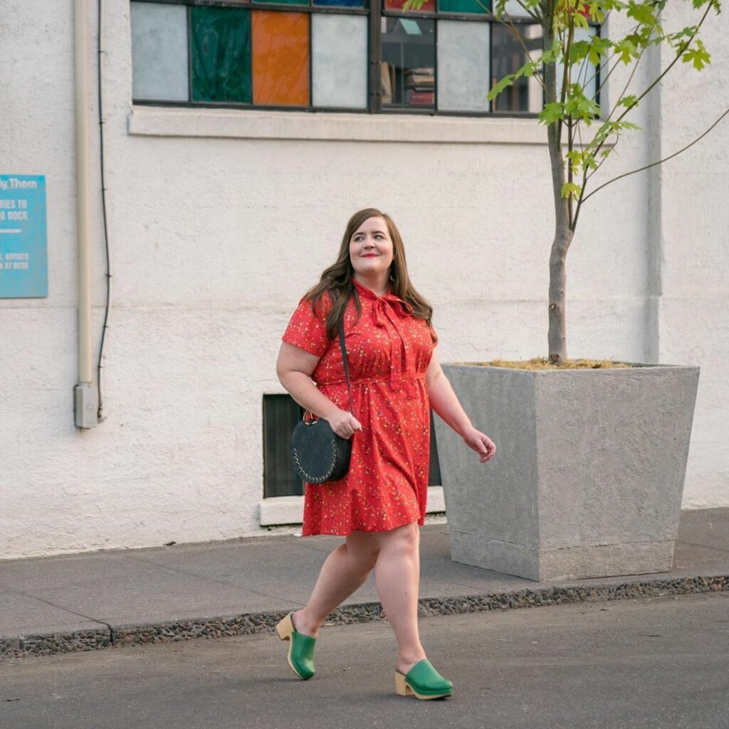 Annie in Shrill. Image: Annie is mid-crossing the street in this photo. She wears mint green clog, a coral flowery short-sleeved dress that is cinched at the waist and ties into a tight collar around her neck. She wears a forest green purse, and looks casually to the left as she crosses the street.