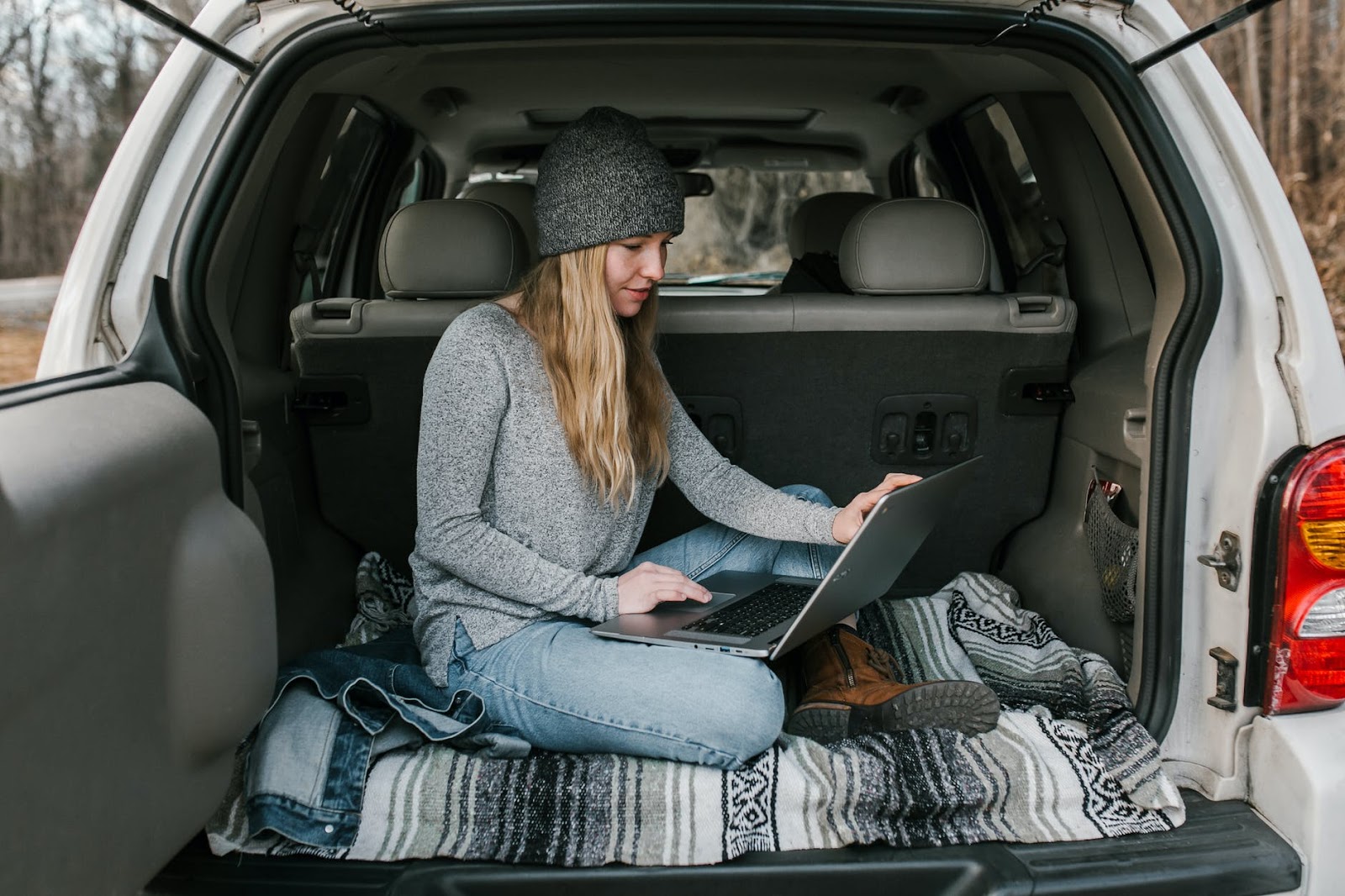 woman in grey sweater and blue denim jeans sitting on a car boot which is open. Lifestyle blogger on open laptop.