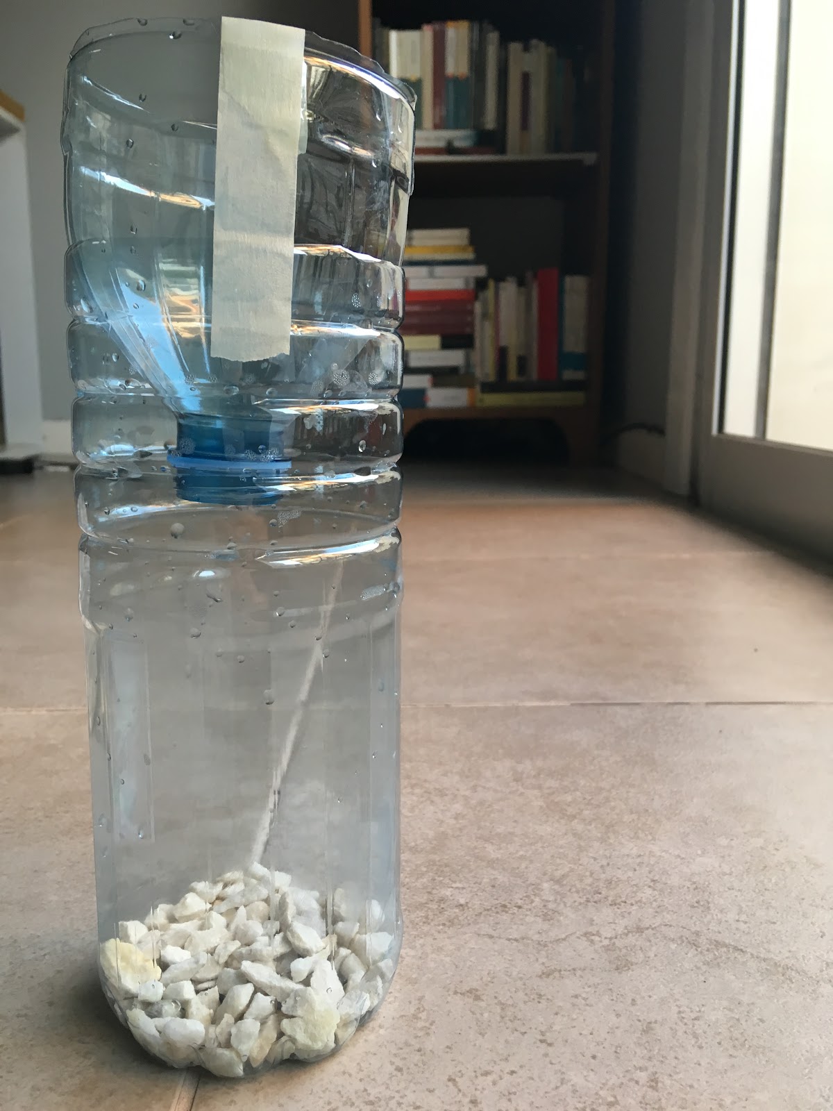 Una botella de plástico cortada con piedras en su base. La sección de la boca de la botella con forma de embudo se encuentra colocada hacia abajo en la botella. 