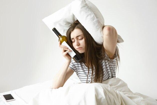 Sad young woman sitting on bed suffering from bad hangover after night party at club, having sleepy tired look, keeping eyes closed, holding bottle of wine and pillow, trying to cover ears from noise Free Photo
