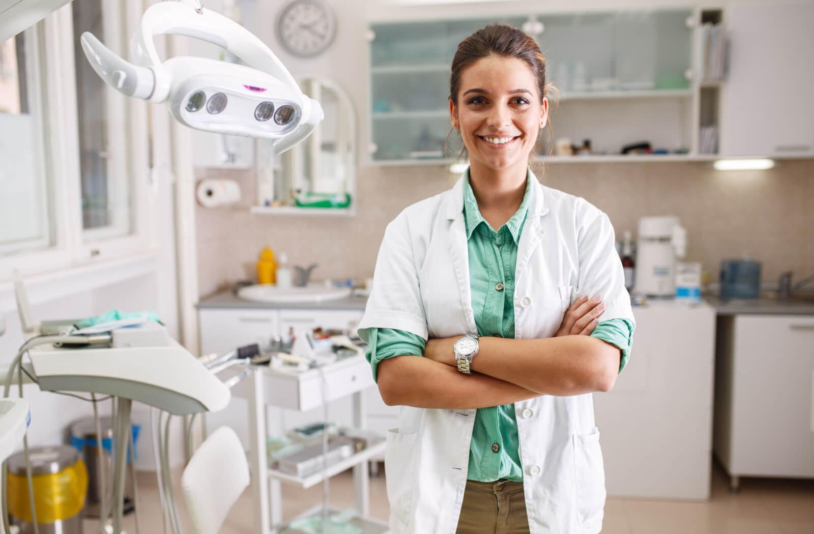 A female dentist smiling and looking directly at the camera with medical equipment in the background.
