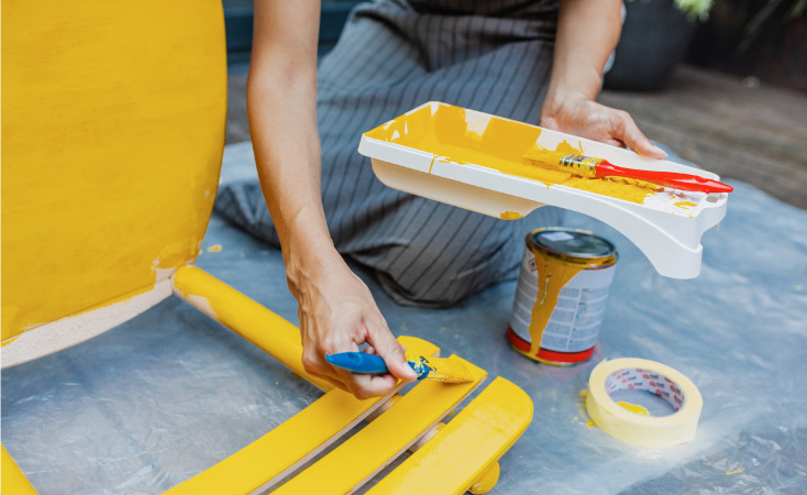 Close-up view of a woman holding a paint tray and using a brush to apply bright yellow paint to the back of a wooden chair. 