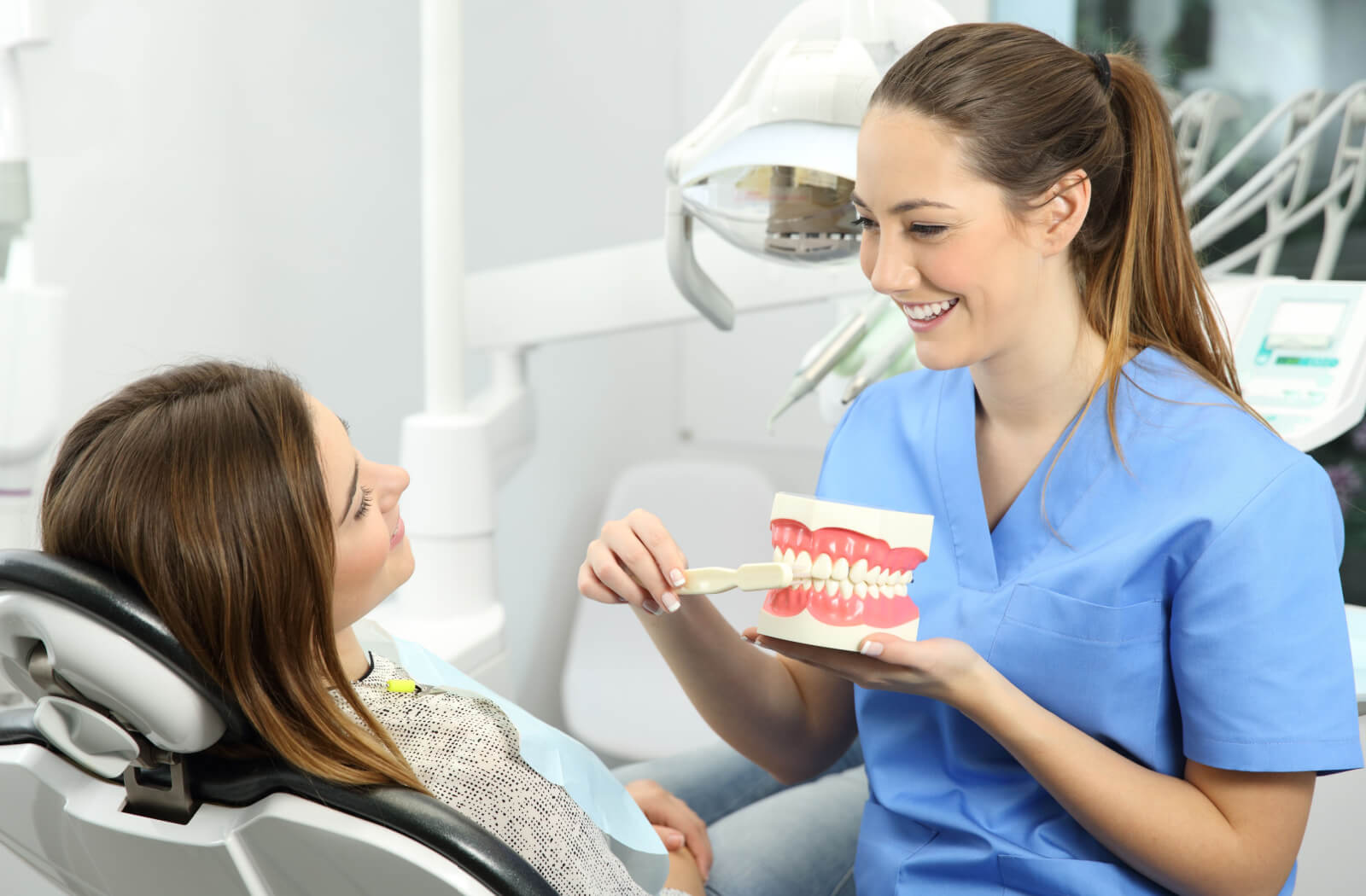A dentist is sitting beside her patient and demonstrating how to properly brush the teeth. Poor oral hygiene can cause gum disease and possible bone loss.