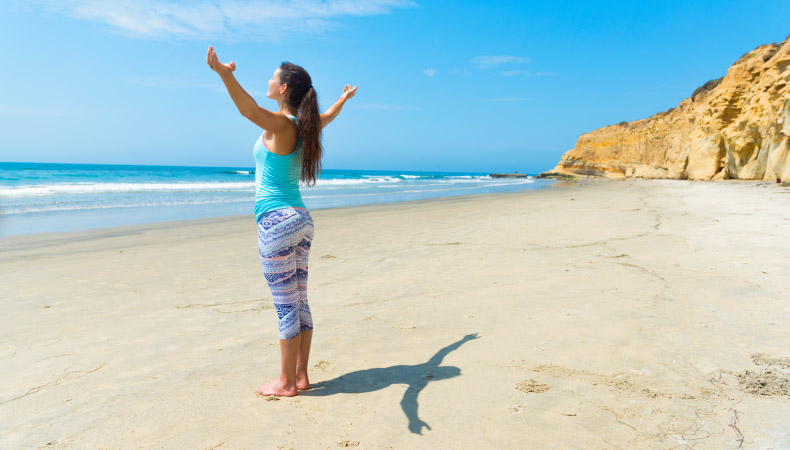 A woman dressed in yoga pants and a tank top stands facing the ocean with her arms outstretched on Black’s Beach in La Jolla, San Diego.