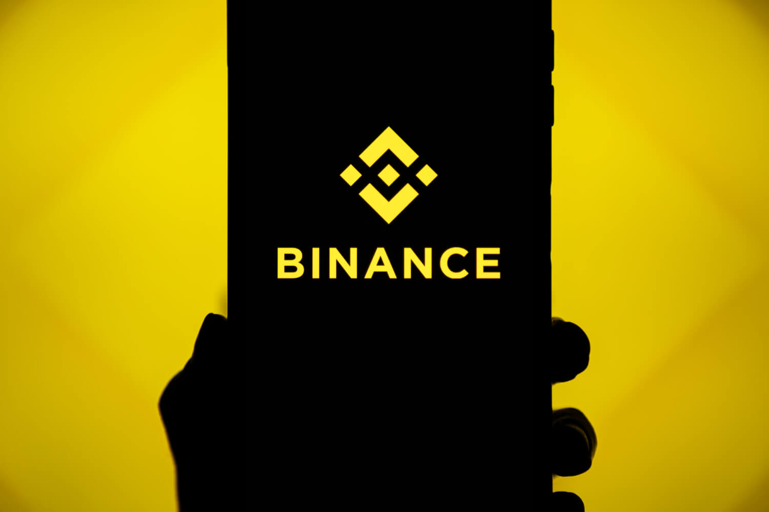 binance logo on a phone held before a yellow backdrop