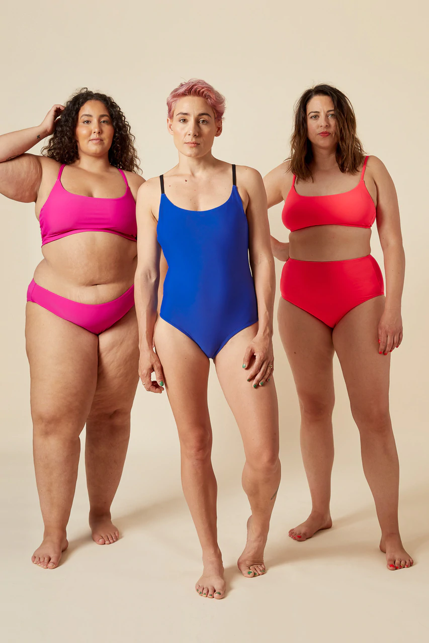Three persons wearing swimsuits.  Person 1 is wearing a hot pink two piece suit with narrow straps & no cups for the top, and a bikini bottom.  Person 2 is wearing a blue one piece suit with narrow straps.  Person three is wearing an orange two piece suit with no cups for the top, narrow straps, and a full brief bottom.
