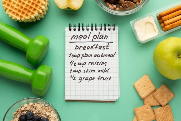 A meal plan notebook that shows the measurements for each recipe, surrounded by fruits, biscuits, waffles, and dumbbells