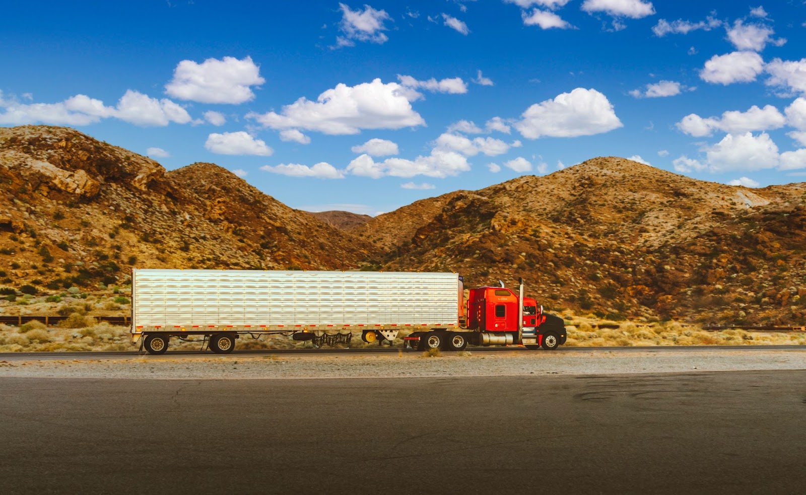 A red semi-truck carrying a load on its back against a backdrop of brown mountains and a blue sky with fluffy white clouds