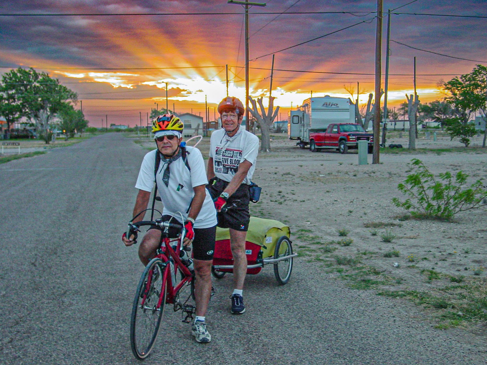 Two cyclists on a tandem bike with a two wheel trailer with sunrise behind them. 