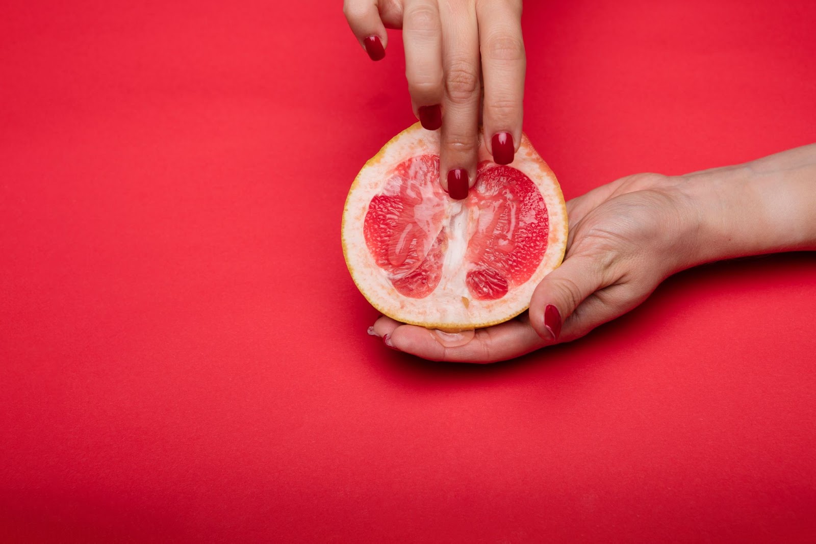 A imagem mostra uma toranja num fundo vermelho. Há a mão de uma mulher com os dedos pressionando a fruta. 
