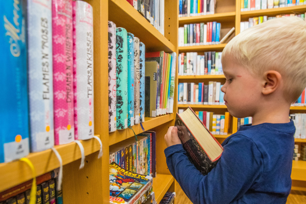 A young boy standing in a library facing a full bookshelf and holding a book.
