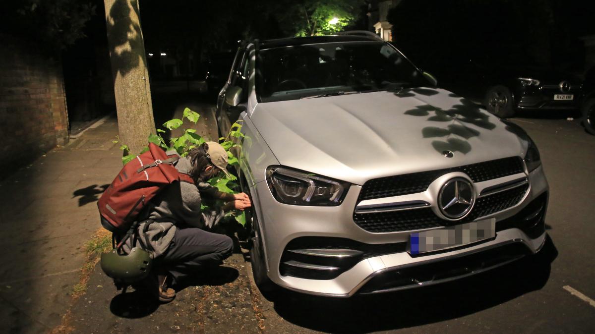 Night-time photo of man with rucksack and some camouflage of twigs with leaves squatting by front tyre of Mercedes car fiddling with air nozzle
