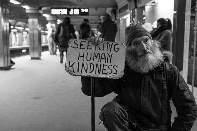 a needy man seated in a train station holding a placard with the words "seeking human kindness"