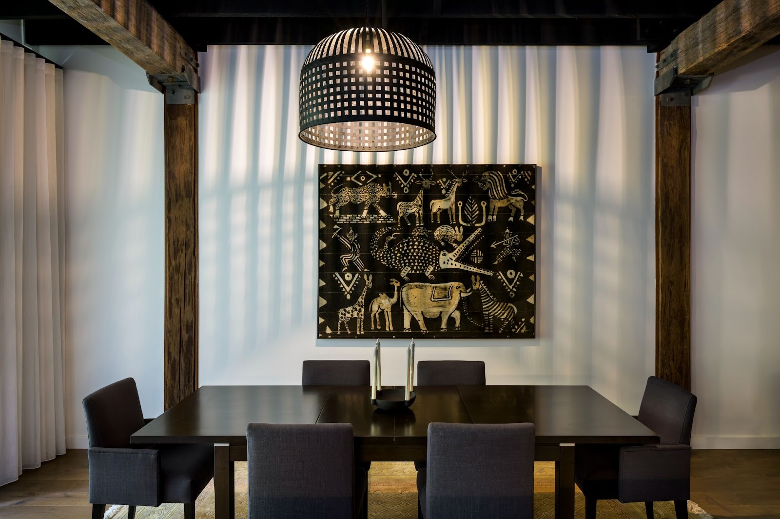 A modern dining room with a large woven pendant light above a dark wooden table. A striking black-and-white artwork with African animal motifs adds a cultural touch to the room, complemented by exposed wooden beams.