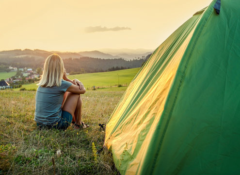 Happy woman with open arms sitting near tent around mountains under sunlight