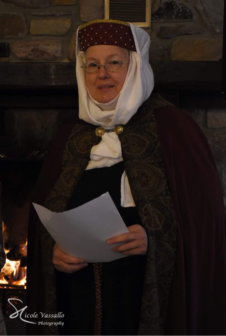 A woman stands facing the camera. She is wearing a white veil over a burgundy hat, as well as a burgundy cloak edged with a gold and blue fabric trim. She is holding a piece of paper