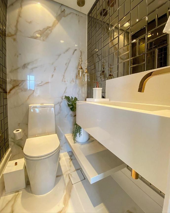 Banheiro com azulejo subway tiles preto na parede da pia e parede lateral, parede de fundo e piso com porcelanato marmorizado em tom claro e  bancada branca com torneira dourada