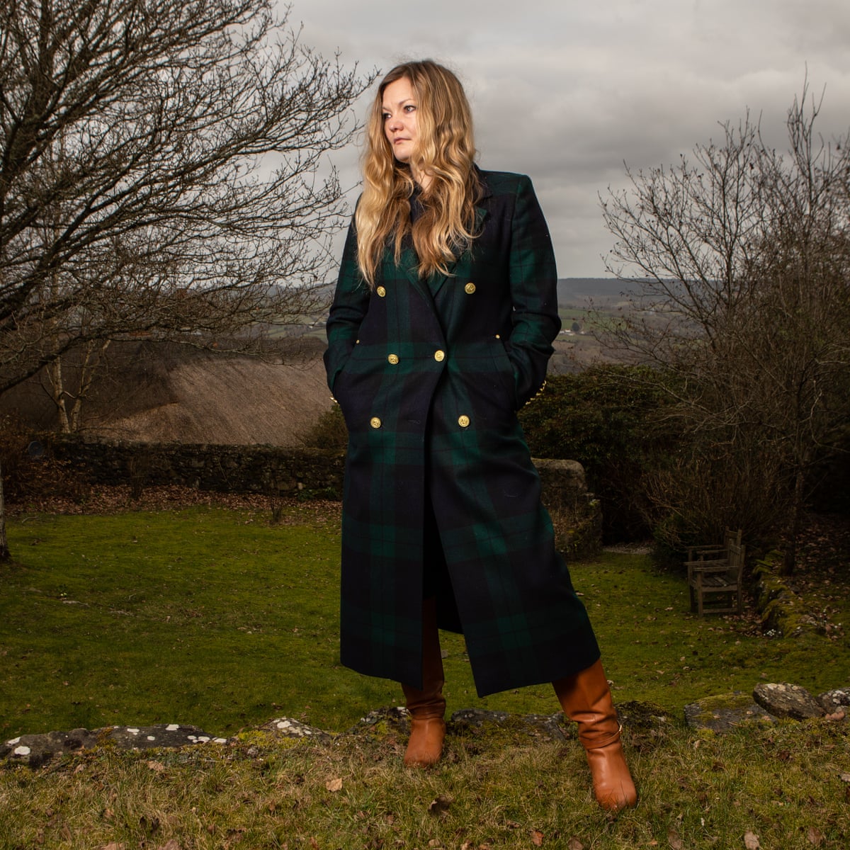This photograph of Catriona Ward features a feminine person with long, wavy blonde hair. Her expression is thoughtful. She is wearing a long green-and-blue plaid coat with wide shoulders and gold buttons, and a pair of tan boots. She stands in front of grey skies over rolling green hills and cliffs.