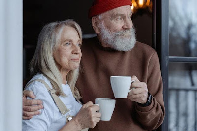 An elderly man and woman standing next to an open window and holding white cups filled with coffee