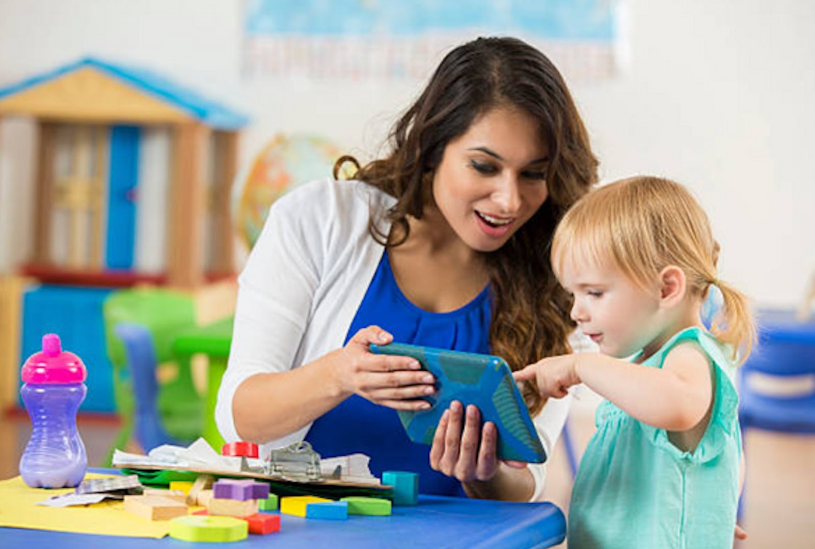 Teacher showing a child pictures on a tablet with different toys and a bottle on the table