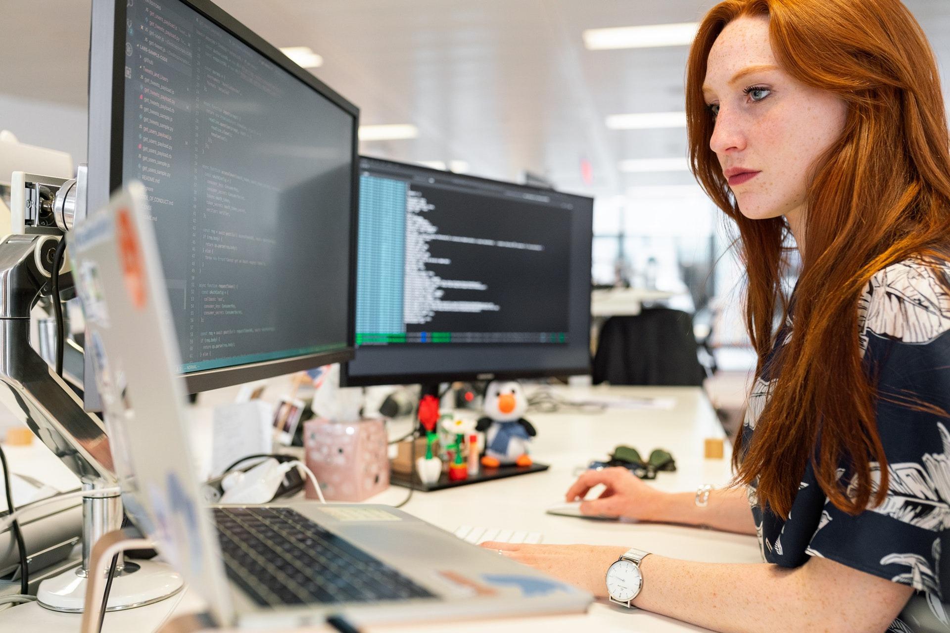 A screem showing a woman working on a computer with two screens.