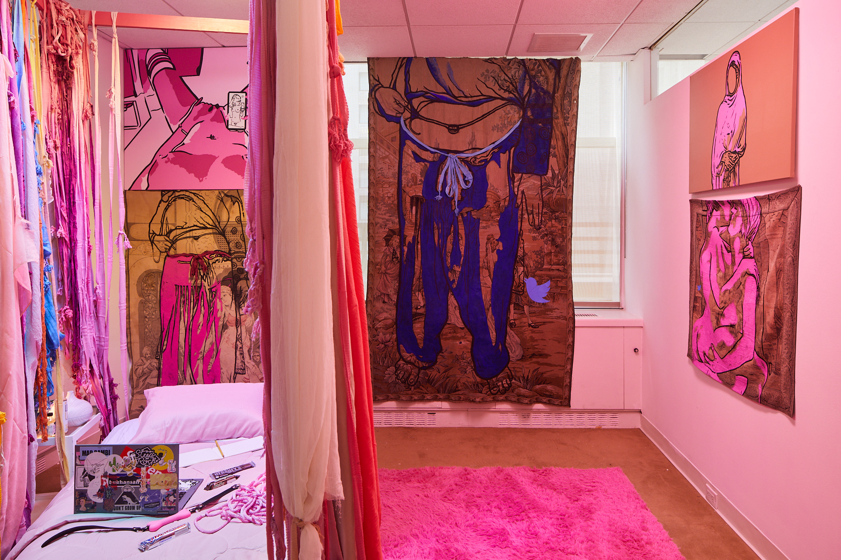 A bright pink room with large hanging images of nude and semi-nude people in erotic poses. Behind the hangings is a low ben with pink sheets, a laptop, and sex toys such as a whip, rope, and a vibrator.