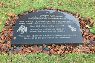 Memorial stone lying on the grass, surrounded by fallen, wet leaves. Carving on the stone dedicates it to the victims of the Essex witch hunts.