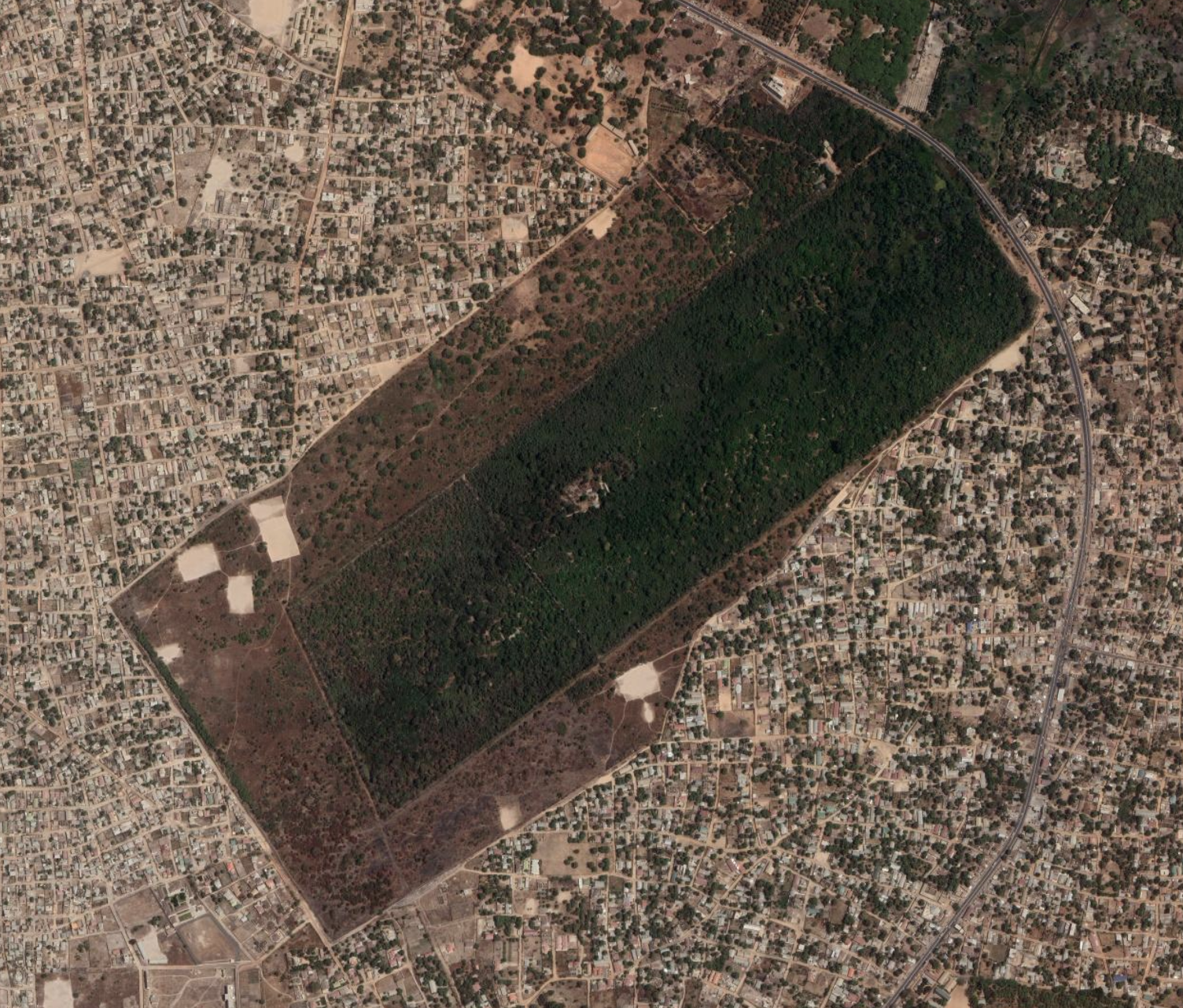 Aerial photograph of Abuko Nature Preserve with a rectangle of dark green, dense forest in the middle, surrounded by brown scrublands, which are surrounded by a dense residential area with minimal green space.