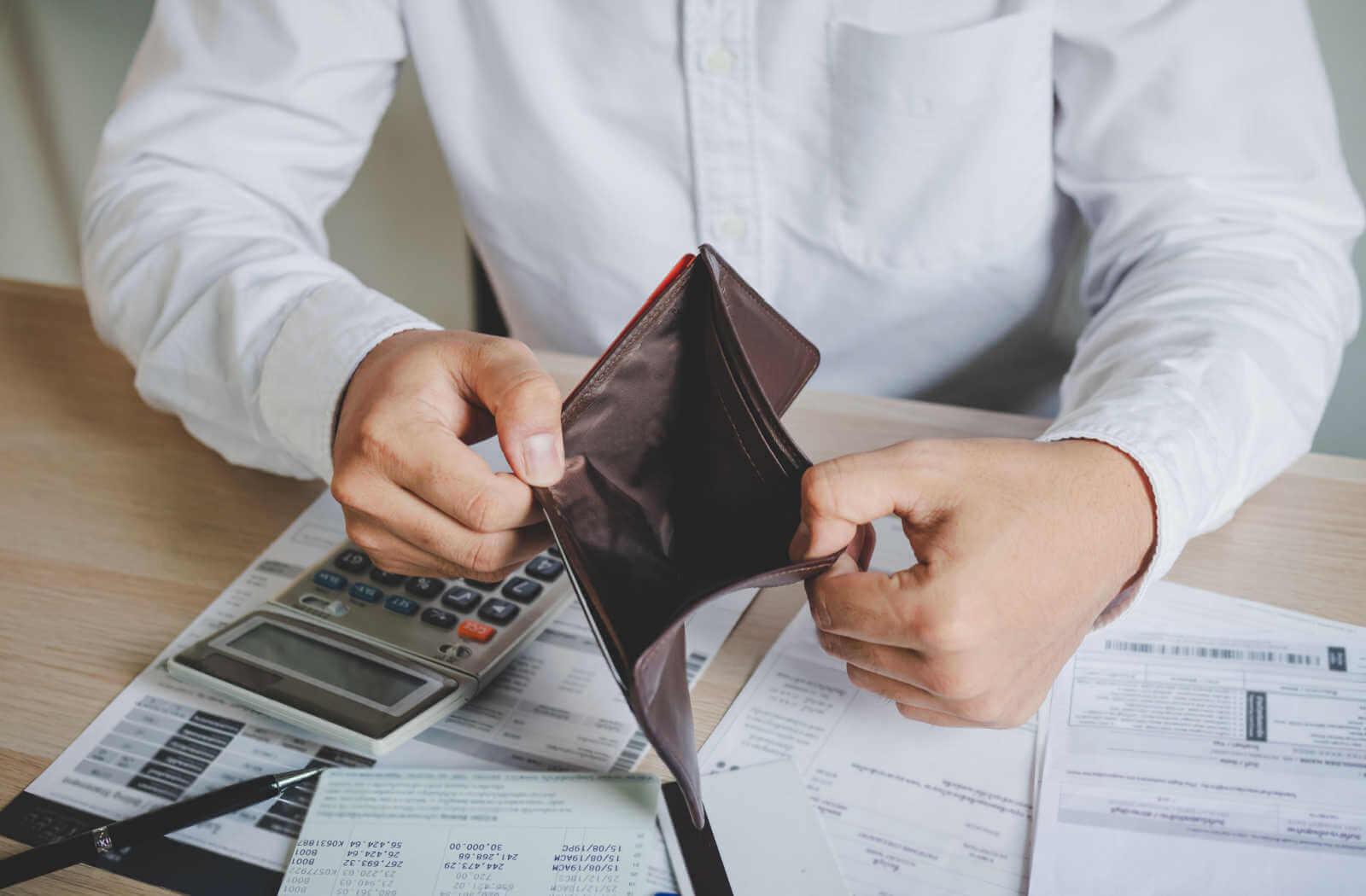 a man sits at a table opening his wallet which is empty. on the table is a calculator and paperwork for filing for bankruptcy