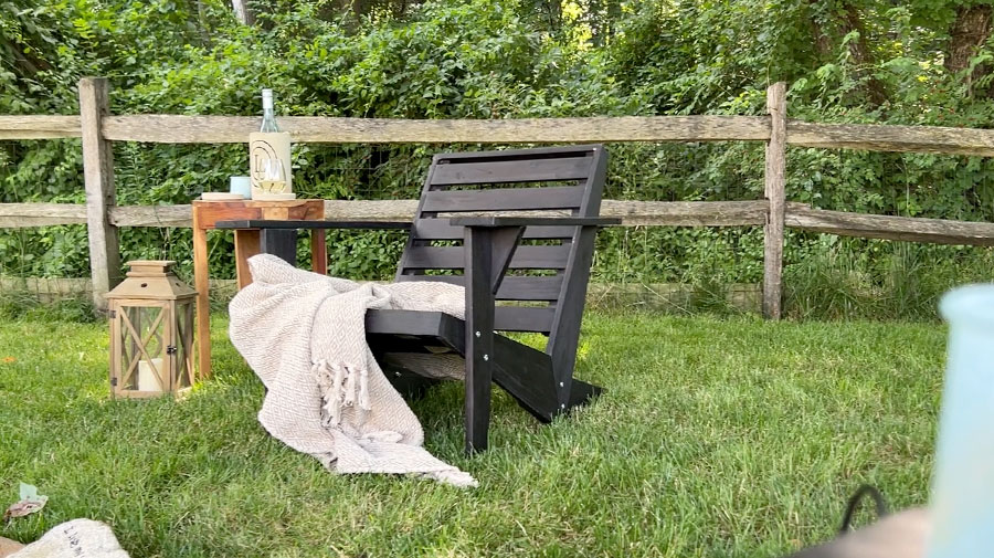 Black Adirondack chair with beige throw next to a side table along a split rail fence
