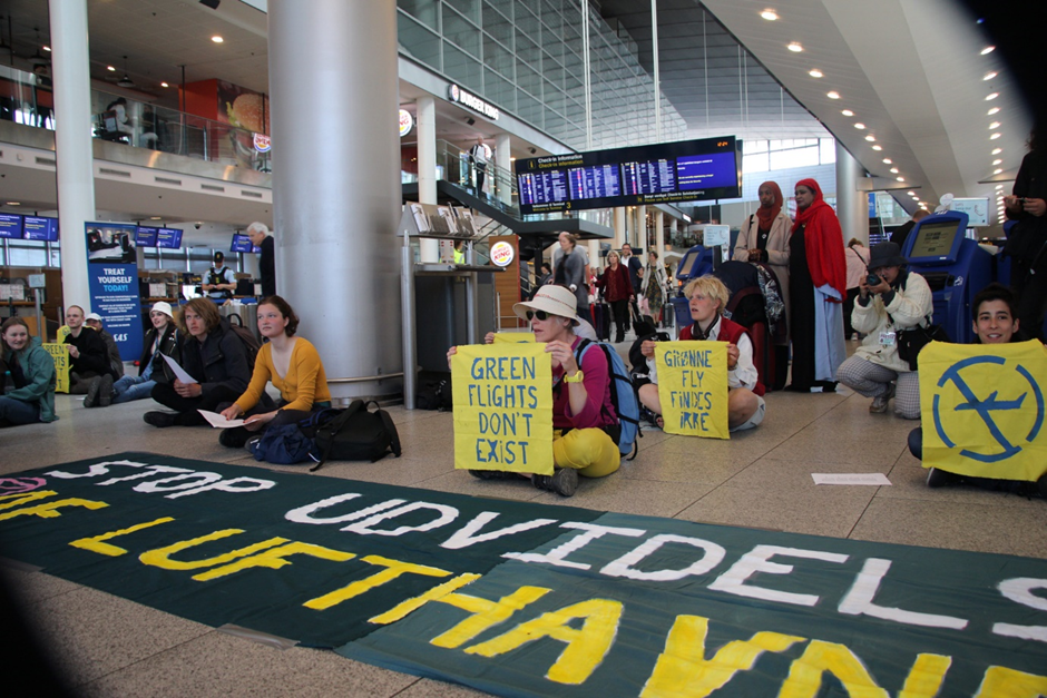 Rebels sit behind a banner on floor of departure lounge holding signs, one saying Green flights don't exist