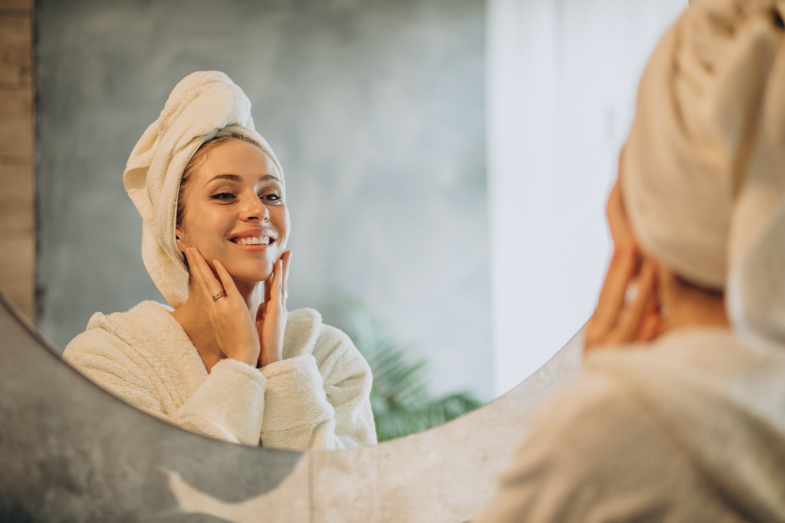 alt text: mulher se olhando no espelho usando um roupão branco e conferindo o resultado da sua skincare 