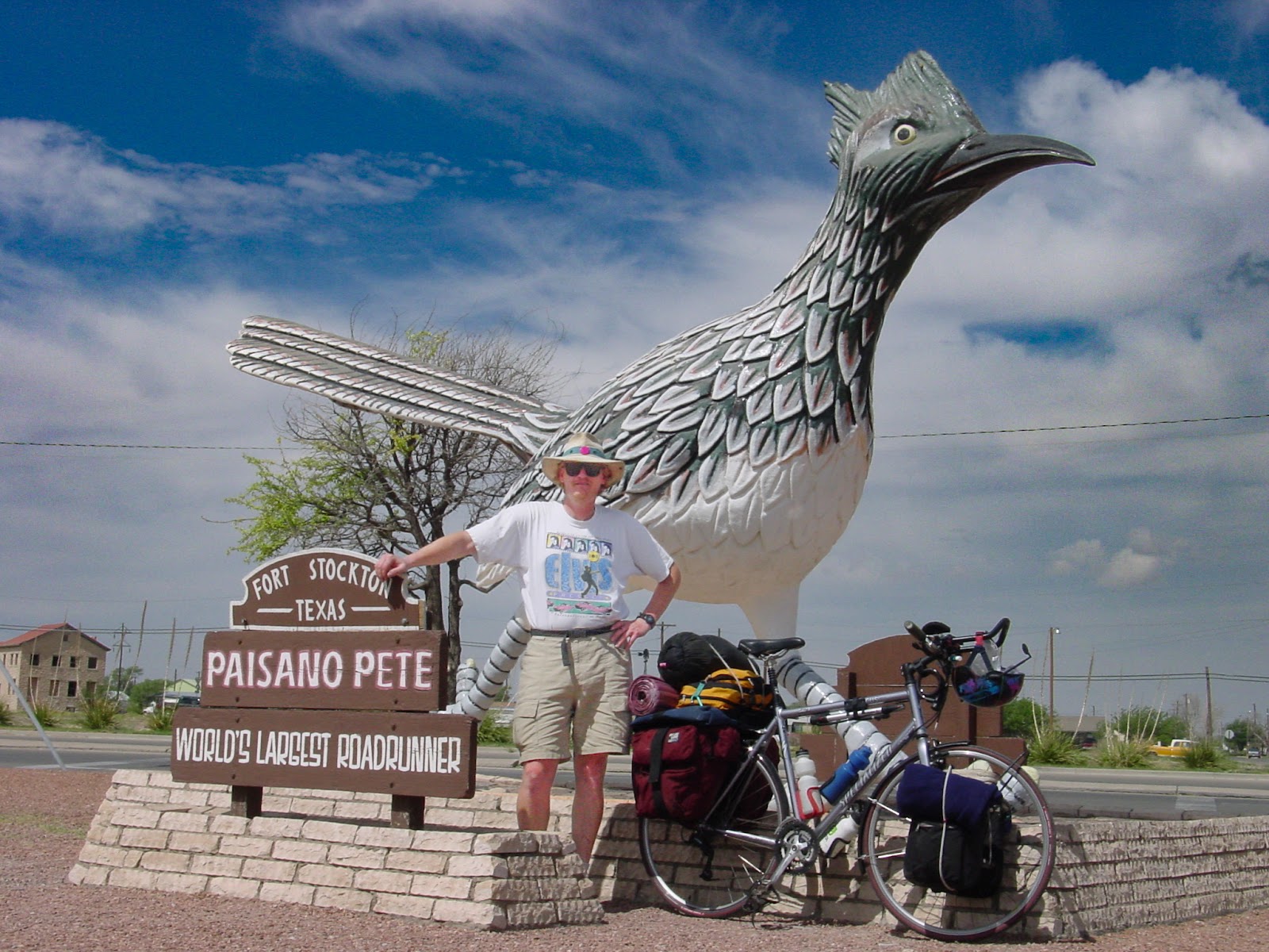 Cyclist stands with bike in front of a two-story tall bird. The sign says: FORT STOCKTON TEXAS, PAISANO PETE WORLD'S LARGEST ROADRUNNER