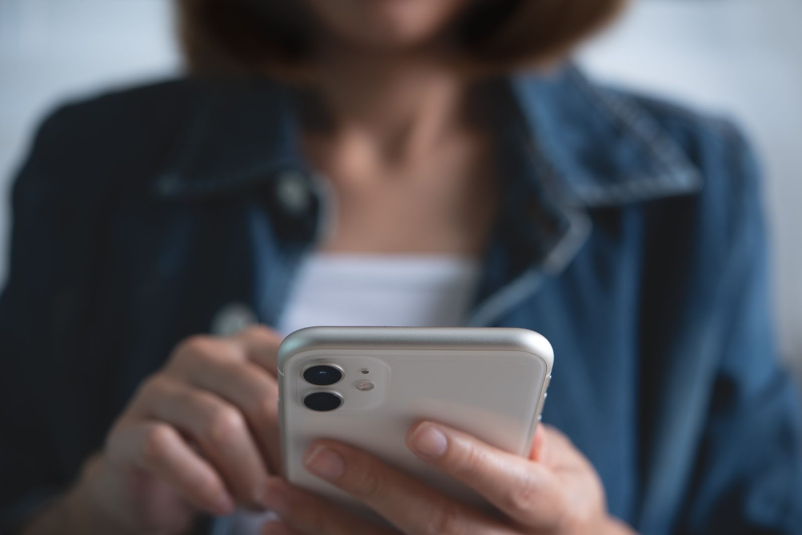 Imagem focada nas mãos de uma mulher segurando e mexendo em seu celular de cor prata. Ao fundo, é possível perceber a imagem do torso desfocada.