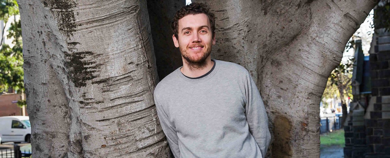 Daniel leans against a huge tree in a church yard and smiles at the camera